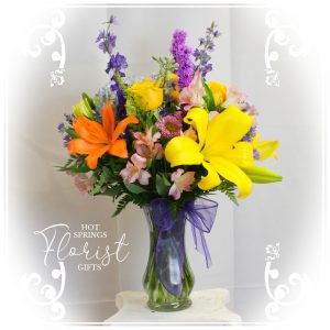 A vibrant bouquet of yellow and orange lilies, purple flowers, and assorted greenery arranged in a clear vase tied with a purple ribbon, displayed against a white background with decorative swirls and a logo for "hot springs florist & gifts".