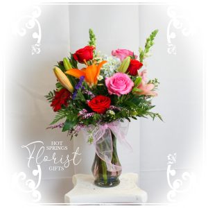 Vibrant mixed flower arrangement in a clear vase with a pink bow, featuring roses and lilies.