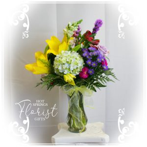 Colorful bouquet of mixed flowers in a clear glass vase on a pedestal with a striped backdrop.