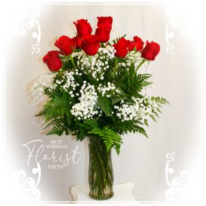 A bouquet of red roses with baby's breath and ferns in a clear vase against a white background.