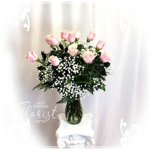 A bouquet of pink roses and baby's breath in a clear vase on a white pedestal with a backdrop of draped fabric.
