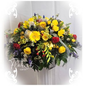 A vibrant floral arrangement with yellow roses, red blooms, and greenery, featuring a sheer ribbon, set against a white backdrop with a floral shop watermark.