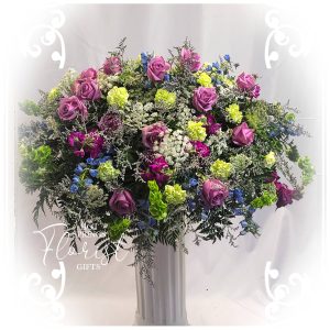 An elegant floral arrangement with a mix of purple roses, blue flowers, greenery, and white accents displayed on a stand.