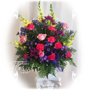 Colorful bouquet of pink roses, purple flowers, and yellow accents arranged in a white pedestal vase against a light background.