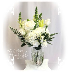 A bouquet of white roses and assorted flowers arranged in a clear vase on a white pedestal with a backdrop including the text "hot springs florist & gifts".