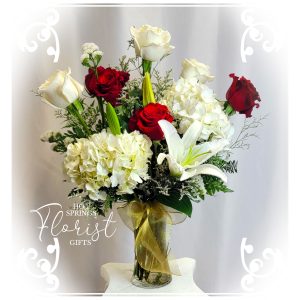 An elegant Holiday Deluxe Seasonal Bouquet (Gold) featuring white lilies, red and white roses, and complementary foliage displayed in a vase with a gold ribbon.