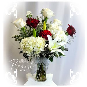 An elegant Holiday Deluxe Seasonal Bouquet (Silver) of white and red flowers, including roses and lilies, arranged in a clear vase on a white pedestal.
