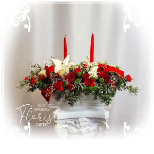 A festive and elegant Holiday Elegant Seasonal Centerpiece with Two Red Candles, white lilies, red roses, and greenery on a white ornamental stand.