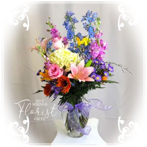 A vibrant floral arrangement featuring a mix of colorful flowers including lilies, delphiniums, and daisies in a glass vase with a purple ribbon, presented on a pedestal.