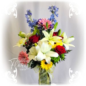 A vibrant bouquet of mixed flowers including lilies, roses, and gerberas displayed elegantly against a gray background with a watermark of "hot springs florist & gifts".