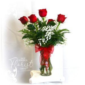 A vase with a bouquet of six red roses and baby's breath flowers, adorned with a red ribbon.