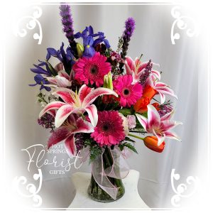 A colorful floral bouquet featuring lilies, gerberas, and other mixed flowers, displayed in a vase with a purple ribbon.