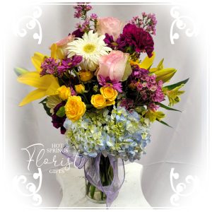 A vibrant Bright Birthday Bundle including roses, lilies, and hydrangeas arranged in a clear vase with a ribbon.