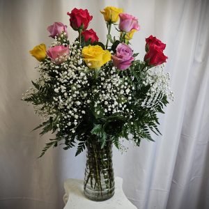 A bouquet of multi-colored roses with baby's breath in a glass vase against a white backdrop.