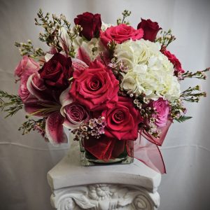 An elegant bouquet of red roses, white flowers, and pink blooms with greenery, displayed on a white pedestal.