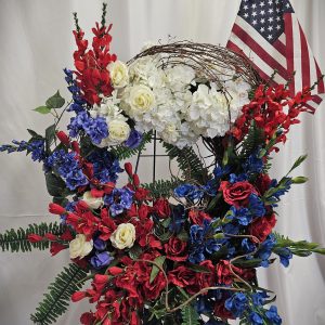 Patriotic floral arrangement featuring red, white, and blue flowers with an american flag in the background.