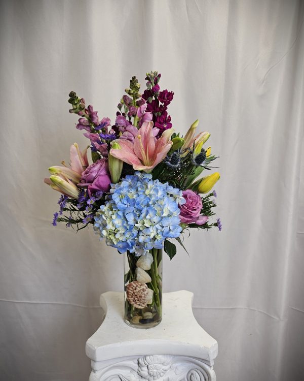 A vibrant bouquet of assorted flowers, including pink lilies and blue hydrangeas, arranged in a glass vase on a white pedestal.