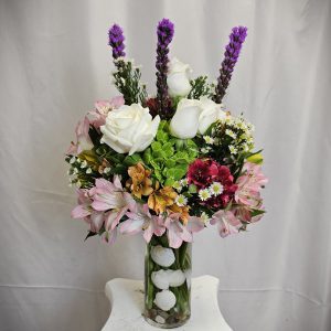 A bouquet of assorted flowers, including white roses and purple spikes, in a clear vase on a white pedestal.