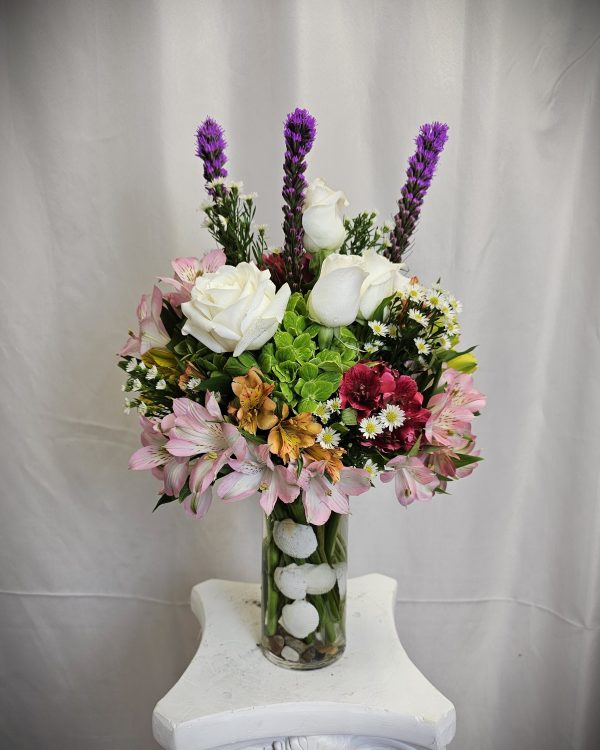 A bouquet of assorted flowers, including white roses and purple spikes, in a clear vase on a white pedestal.
