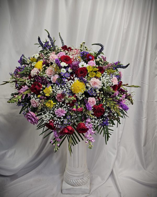 A large, colorful floral arrangement on a white pedestal with a draped background.