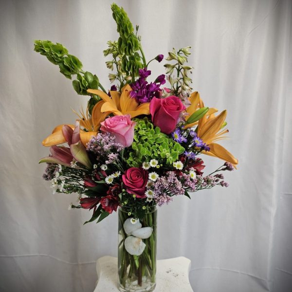 A vibrant bouquet with roses, lilies, and assorted flowers in a glass vase against a white backdrop.