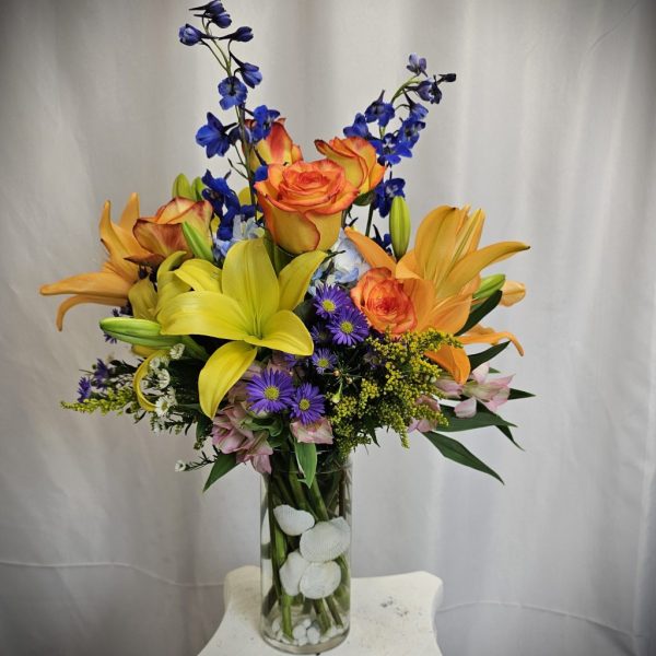 Vibrant floral arrangement featuring lilies, roses, and delphinium in a clear glass vase.