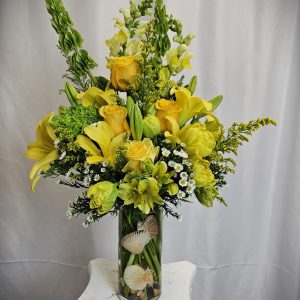 A vibrant floral arrangement with yellow lilies, white flowers, and greenery in a glass vase adorned with seashells.