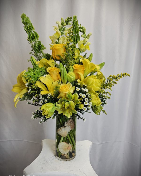 A vibrant floral arrangement with yellow lilies, white flowers, and greenery in a glass vase adorned with seashells.