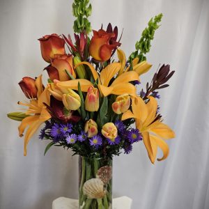 A vibrant floral arrangement featuring orange lilies, red and yellow tulips, and purple accents, presented in a clear vase with seashells at the base.