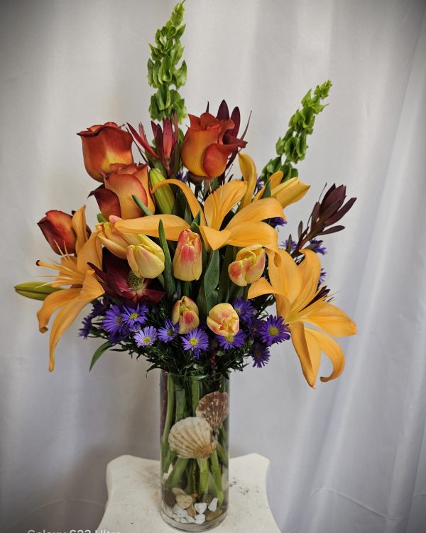 A vibrant floral arrangement featuring orange lilies, red and yellow tulips, and purple accents, presented in a clear vase with seashells at the base.