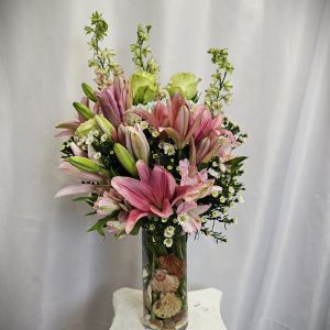 A bouquet of pink lilies, greenery, and white flowers arranged in a clear glass vase on a white pedestal.