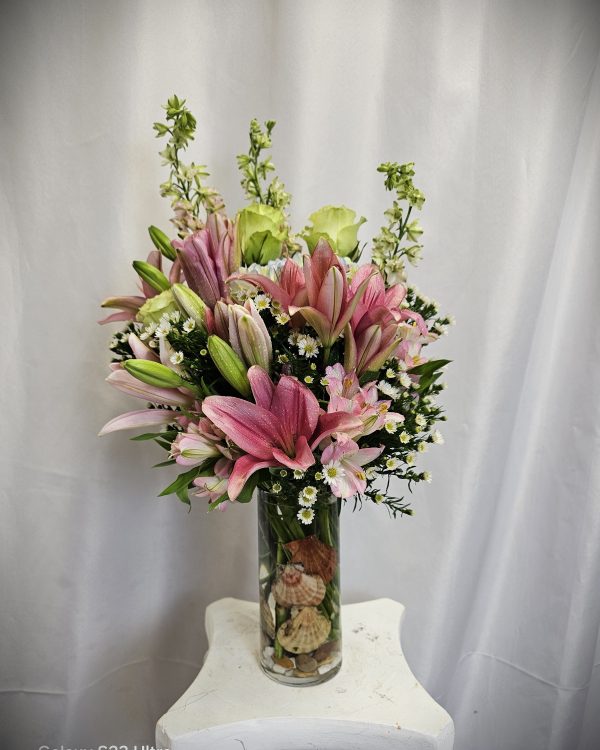 A bouquet of pink lilies, greenery, and white flowers arranged in a clear glass vase on a white pedestal.