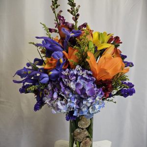 A vibrant bouquet of mixed flowers, including lilies, irises, and hydrangeas, arranged in a glass vase on a pedestal.