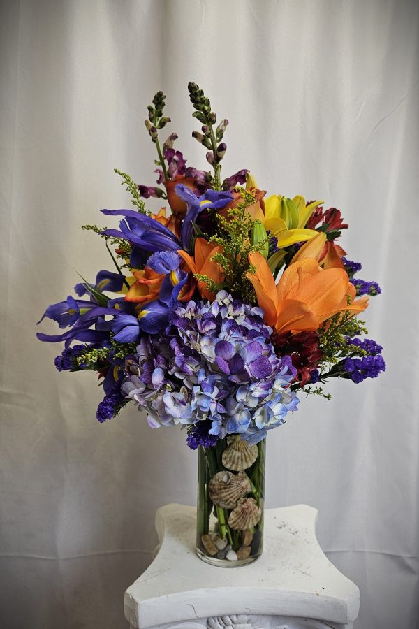 A vibrant bouquet of mixed flowers, including lilies, irises, and hydrangeas, arranged in a glass vase on a pedestal.