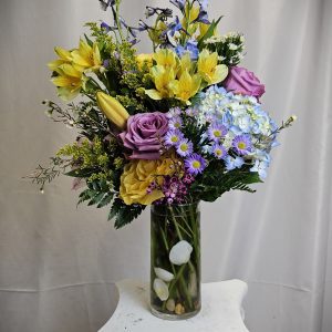 A vibrant floral arrangement featuring yellow lilies, purple roses, and blue delphiniums in a clear vase.