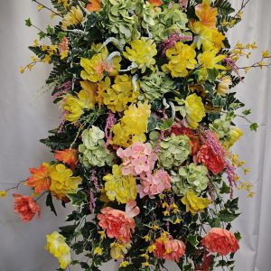 A large floral arrangement featuring a variety of colorful artificial flowers, including yellow, pink, and peach blossoms, with green foliage and small yellow accents, displayed against a light background—perfect as funeral flowers to convey heartfelt condolences.