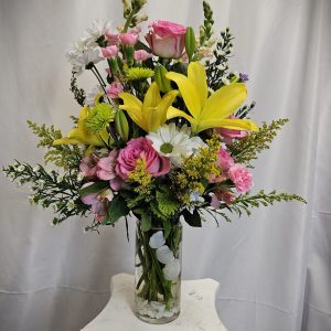 A tall glass vase filled with an arrangement of pink roses, yellow lilies, white daisies, and various greenery, placed on a white pedestal against a neutral background.