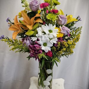 A tall glass vase contains a colorful arrangement of flowers, including white daisies, yellow lilies, and various others, set against a neutral backdrop.