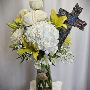 A floral arrangement with white roses, white hydrangeas, and yellow lilies in a glass vase containing decorative rocks and shells, adorned with an American flag themed cross.