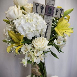 A floral arrangement of white roses, hydrangeas, and yellow lilies in a vase, adorned with a cross inscribed with "And I will give unto thee the keys of the kingdom of heaven.