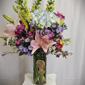 A glass vase filled with a variety of flowers including pink lilies, purple roses, yellow snapdragons, and blue hydrangeas, with seashells placed at the base.