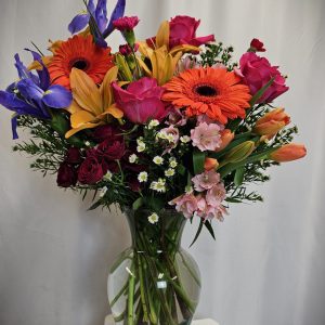 A vibrant Garden Vase of mixed flowers including gerberas, roses, and lilies in a glass vase, set against a plain white backdrop.