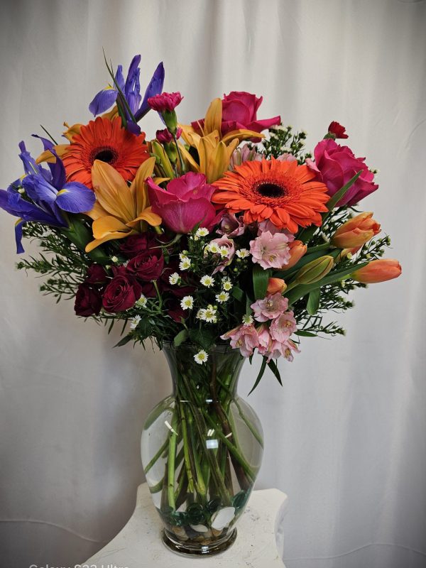 A vibrant Garden Vase of mixed flowers including gerberas, roses, and lilies in a glass vase, set against a plain white backdrop.