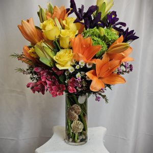 A vibrant bouquet of Orange Asiatic and Yellow Roses, arranged in a clear vase on a white pedestal.