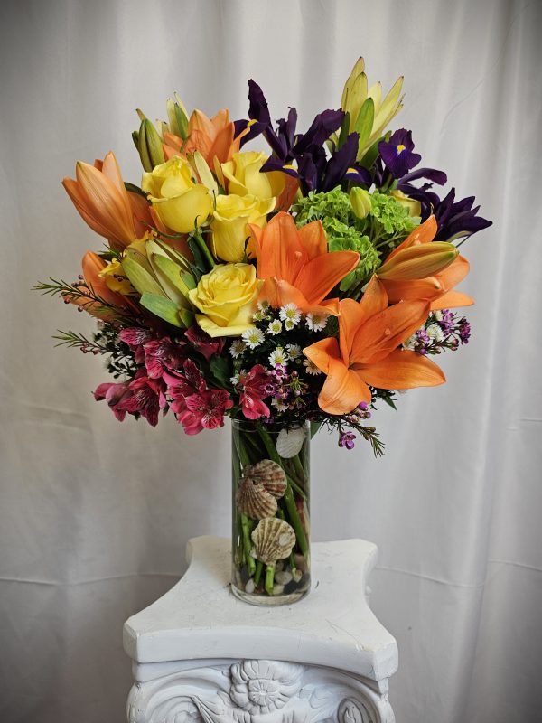 A vibrant bouquet of Orange Asiatic and Yellow Roses, arranged in a clear vase on a white pedestal.