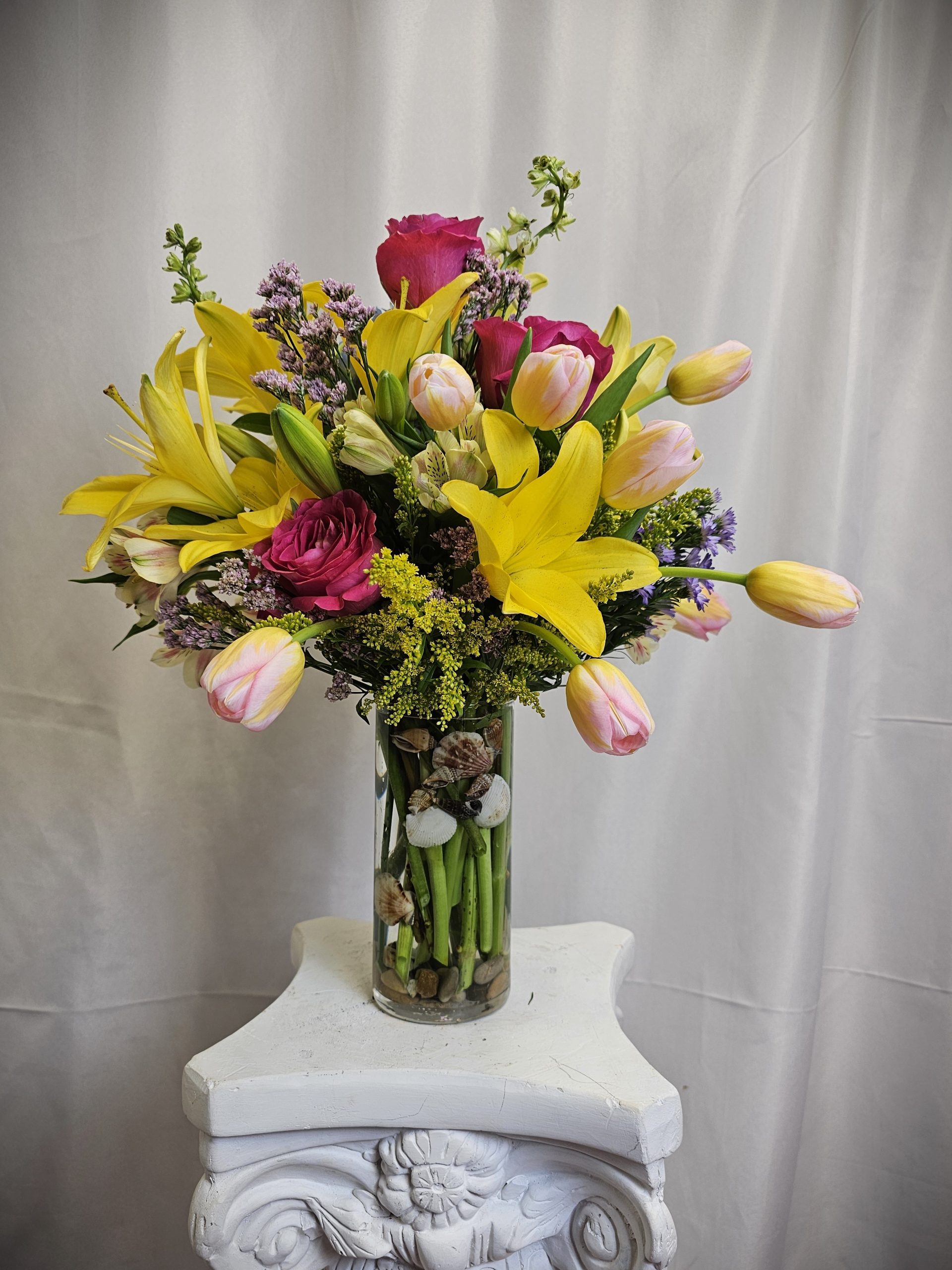A vibrant bouquet of Orange Tulips and Purple Iris' in a glass vase on a white pedestal.