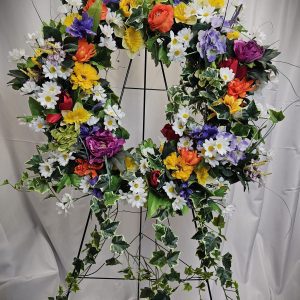 A wreath made of colorful artificial funeral flowers, including daisies, roses, and ivy leaves, displayed on a black metal stand with a white fabric backdrop.