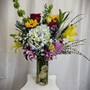 A tall glass vase holds an arrangement of colorful flowers, including sunflowers, lilies, hydrangeas, and roses, with seashells at the base, standing on a white pedestal against a white backdrop.