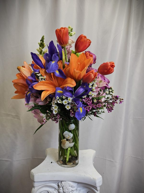A vibrant bouquet of Orange Tulips and Purple Iris' arranged in a glass vase on a white pedestal.