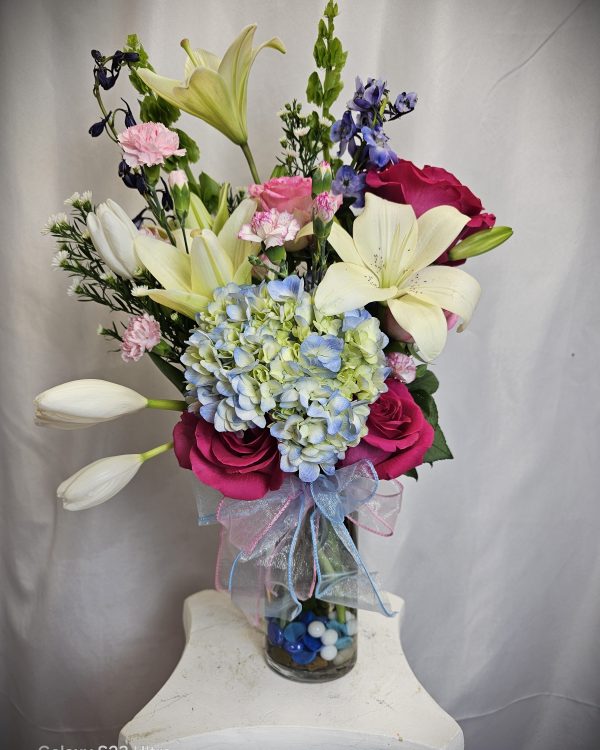 A bouquet of mixed flowers including lilies, roses, hydrangeas, and carnations arranged in a glass vase filled with decorative stones. The vase sits on a white pedestal against a plain backdrop.